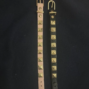 Armband med nitar i guld, 2-pack - Två smala armband i läder, ett i ljusrosa och ett i svart, med coola fyrkantiga guldfärgade nitar längs hela bandet. Båda har guldfärgat spänne och detaljer. Perfekt accessoar för att ge din outfit en edgy vibe.
