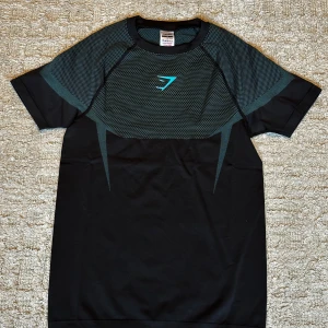 Gymshark Onyx m - Snygg svart t-shirt från Gymshark med coola meshdetaljer över axlar och bröst. T-shirten har korta ärmar, sportig passform och en blå Gymshark-logga på bröstet. Perfekt för dig som gillar att träna och vill ha en stilren look på gymmet.