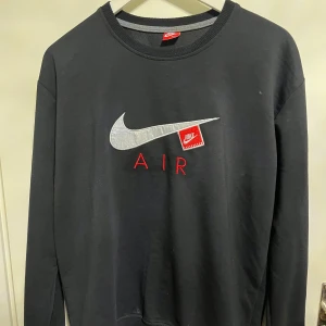 Svart Nike Air sweatshirt med broderi - Svart sweatshirt från Nike med stort silverfärgat swoosh och röd broderad Air-text på bröstet. Tröjan har rund hals och långa ärmar, perfekt för en chill och sportig look. Klassisk Nike Air-design med snygga detaljer.