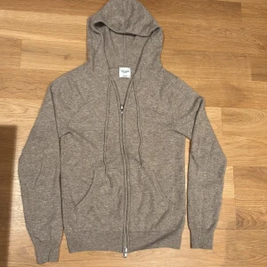 Beige cashmere hoodie från John Henric - Mysig beige hoodie i 100% kashmir från John Henric. Tröjan har dragkedja framtill, huva med snörning och två fickor. Perfekt för dig som vill ha en lyxig och stilren look med maximal komfort.