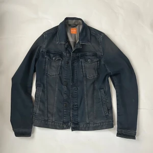Mörkblå jeansjacka från BOSS - Snygg mörkblå jeansjacka från BOSS med klassisk krage, knappar framtill och två bröstfickor med lock. Jackan har långa ärmar och coola söm-detaljer som ger en schysst streetkänsla. Perfekt för dig som gillar stilren och tidlös design.