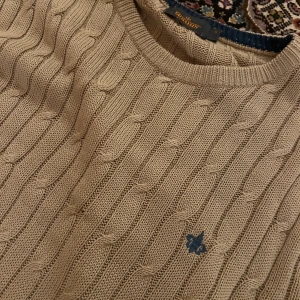 Beige stickad tröja från Morris - Säljer en beige kabelstickad tröja från Morris med rund halsringning och diskret logga broderad på bröstet. Tröjan har lång ärm och är gjord i mjuk bomull, perfekt för lager-på-lager under höst och vinter.