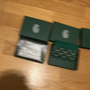 Gröna smyckesaskar med logotyp - Två eleganta gröna smyckesaskar med silverfärgad logotyp på locket. Perfekta för att förvara eller ge bort smycken som armband, halsband eller örhängen. Askarna har en lyxig känsla och insidan är vadderad för extra skydd.