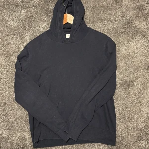 Svart hoodie från Grunt i storlek L - Svart hoodie från Grunt i storlek xs. Tröjan har en klassisk huva och är långärmad med ribbade muddar vid ärmslut och nederkant.  Materialet är merinoull , vilket gör den skön att bära. Perfekt för dig som gillar enkel och stilren design. Den har ett pytte litet hål i armhålan som kanske är runt en halv centimeter stort😁