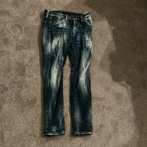 Blå jeans från Jack & Jones - Säljer ett par blå jeans från Jack & Jones med coolt slitna detaljer och ljusa tvättade partier. Jeansen har raka ben, klassiska fickor och tydliga sömmar. Perfekt för dig som gillar en avslappnad streetstil. 30-32 size som sitter slimfit med slitningar