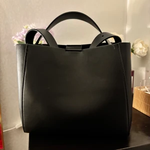 Svart stilren handväska i skinn - Snygg svart handväska i minimalistisk design med stilrena linjer och två handtag. Väskan är tillverkad i slätt skinn och har en boxig form som ger ett modernt intryck. Perfekt för dig som gillar en clean och klassisk look.