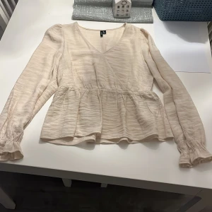 Beige blus från Vero Moda💕 - Säljer en ljusbeige blus från Vero Moda i storlek S. Blusen har omlottdesign med v-ringning, volang vid midjan och långa ärmar med rynkade muddar. Materialet är lätt och har en strukturerad yta som ger en snygg touch. Perfekt för dig som gillar romantiska detaljer.  Prislappen är av men tröjan är inte använd! Skriv om du undrar något eller vill ha fler bilder💕
