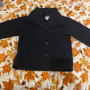 Svart rock från Gina Tricot - Svart rock från Gina Tricot med klassisk krage och tre knappar framtill. Jackan har en stilren och rak passform, perfekt för dig som gillar enkel och clean stil. Materialet känns mjukt och har en snygg struktur. Passar dig som vill ha en tidlös look.