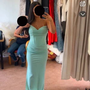 Ljusgrön långklänning med snörning - Säljer en elegant ljusgrön långklänning med v-ringad och axelbandslös design. Klänningen har snygga draperingar framtill och snörning i ryggen för en perfekt passform. Materialet känns mjukt och har ett lätt glansigt utseende, vilket ger en lyxig känsla. Extra kostnad lades på för skräddaranpassningen.