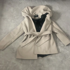 Beige kappa med bälte Classic Collection - Snygg beige kappa från Classic Collection i storlek 38. Jackan har en stor krage, bälte i midjan och knappar på insidan. Materialet är mjukt och känns lyxigt, perfekt för dig som gillar stilrena och eleganta plagg. Fodrad med svart tyg på insidan.