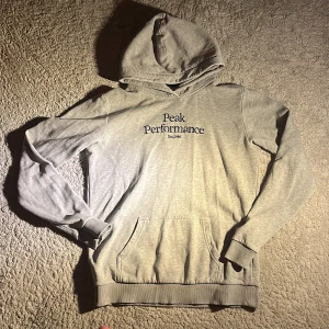 Grå hoodie från Peak Performance - Säljer en grå hoodie från Peak Performance med broderad logga på bröstet. Tröjan har huva, magficka och ribbade muddar vid ärmslut och nederkant. Perfekt för en avslappnad stil.