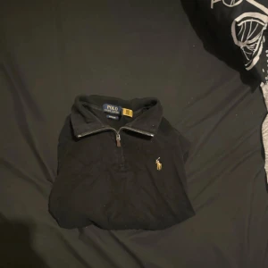 Svart half zip tröja från Polo Ralph Lauren - Äkta finns bevis  Svart långärmad tröja från Polo Ralph Lauren med klassisk broderad logga på bröstet. Tröjan har en half zip-dragkedja vid kragen och är gjord i 100% bomull. Perfekt för dig som gillar stilrena och tidlösa plagg.
