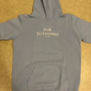 Ljusblå hoodie Peak Performance - Snygg ljusblå hoodie från Peak Performance med vit logga på bröstet. Tröjan har huva, magficka och ribbade muddar vid ärmslut och nederkant. Perfekt för chill dagar och enkel att matcha med jeans eller mjukisbyxor.