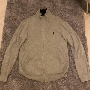 Grå kofta från Polo Ralph Lauren - Snygg grå kofta från Polo Ralph Lauren med hel dragkedja och klassisk ståkrage. Tröjan har långärmad passform, två fickor fram och den ikoniska Polo-loggan broderad på bröstet. Tillverkad i mjuk bomull som känns skön mot huden.