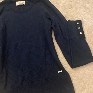 Marinblå stickad tröja Holly & Whyte - Snygg marinblå stickad tröja från Holly & Whyte by Lindex i storlek XS. Tröjan har långärmade ribbade muddar med tre dekorativa guldfärgade knappar och en liten metallbricka vid nederkanten. Perfekt för dig som gillar stilrena detaljer.