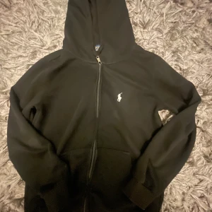 Svart hoodie från Polo Ralph Lauren - Svart hoodie från Polo Ralph Lauren med dragkedja framtill och klassisk vit broderad logga på bröstet. Tröjan har huva, långa ärmar och känguruficka. Perfekt för en chill och stilren look. Den passar för typ 160-163