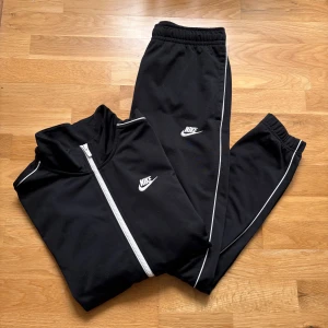Svart Nike set - Svart set från Nike med ziptröja och tillhörande byxor. Tröjan har hel dragkedja framtill, hög krage och smala vita ränder längs ärmarna. 
