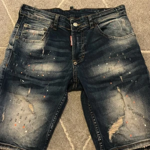 Dsquared2 jeansshorts med slitningar - Snygga jeansshorts från Dsquared2 i mörkblå denim med slitna detaljer och färgstänk i vitt och orange. Klassisk femficksmodell med gylf och knapp, samt Dsquared2-logga på etikett och bakficka. Perfekt för dig som gillar streetstyle och edgy look. (Inte äkta) små i storleken 
