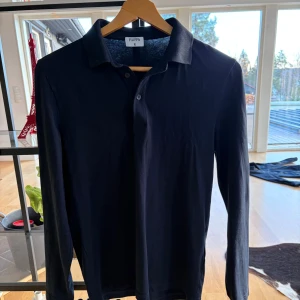 Mörkblå pikétröja från Filippa K - Snygg mörkblå långärmad pikétröja från Filippa K. Klassisk krage och knäppning med två knappar framtill. Tillverkad i mjuk bomull som känns skön mot huden. Perfekt för en stilren och avslappnad look.