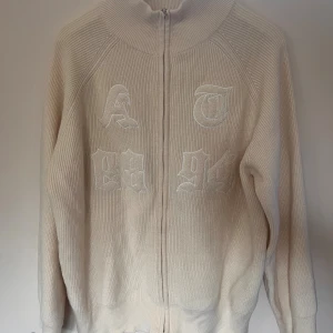 Beige stickad kofta från Dazy - Beige stickad kofta från Dazy med dragkedja framtill och broderade bokstäver och siffror i vitt på bröstet. Tröjan har hög krage och långa ärmar, perfekt för lager-på-lager. Materialet är 100% akryl och modellen är normal i passformen.