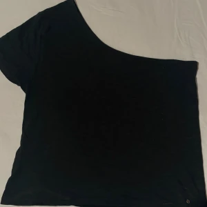 Svart oneshoulder topp från Brownie - Svart topp från Brownie i oneshoulder-modell med kort ärm på ena sidan. Tunn och mjuk, perfekt för dig som gillar minimalistisk stil. Materialet känns som bomull och toppen har en clean look utan mönster eller detaljer. Snygg att matcha med jeans eller kjol.