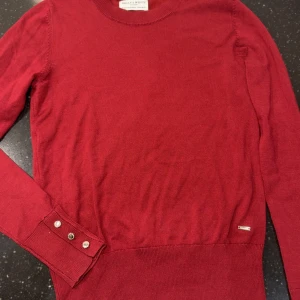 Röd stickad tröja Holly & Whyte XS - Snygg röd stickad tröja från Holly & Whyte by Lindex i storlek XS. Tröjan har rund hals, långa ärmar med dekorativa knappar vid mudden och en liten metallbricka vid nederkanten. Perfekt för dig som gillar klassisk stil med en modern touch. Den ser mer vinröd ut i verkligheten, ljuset gör det mer ljusare 