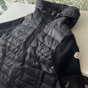 Svart pufferjacka från Moncler - Snygg svart pufferjacka från Moncler med quiltad framsida och huva med snörning. Jackan har dragkedja framtill, ribbade ärmar och Moncler-logga på armen. Perfekt för dig som gillar stilren och sportig look.