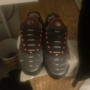 Nike TN svarta och röda sneakers - Nike TN sneakers i svart med röda och vita detaljer. Snyggt mönstrad ovandel med vågiga linjer, röd Swoosh på sidan och Tn-logga bak. Platt sula med synlig Air-enhet och snörning framtill. Insidan är röd och skorna har en sportig vibe.