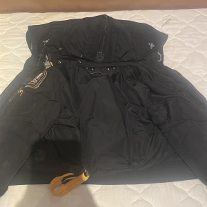 Svart Parajumpers dunjacka med fuskpäls - Säljer en svart Parajumpers dunjacka i slim fit med snygga detaljer som patch på ärmen, orange rem och fuskpäls i huvan. Jackan har flera fickor, dragkedja och är perfekt för kalla vinterdagar. Klassisk och sportig look med robust känsla.      ⚠️OBS ⚠️ utre päls krage saknas!