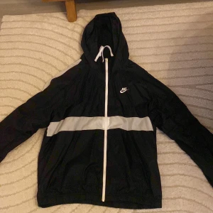 Svart vindjacka från Nike med huva - Svart vindjacka från Nike med vit dragkedja, huva och en bred vit rand över bröstet och axlarna. Jackan har lång ärm och är tillverkad i lätt polyester, perfekt för blåsiga dagar. Klassisk Nike-logga på bröstet och elastiska muddar nedtill.