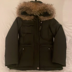 Svart vinterjacka med päls från Hollies - Snygg svart vinterjacka från Hollies med stor huva och fluffig pälsimitation runt kanten. Jackan har flera fickor framtill, dragkedja och knappar. Perfekt för kalla dagar och riktigt stilren look.