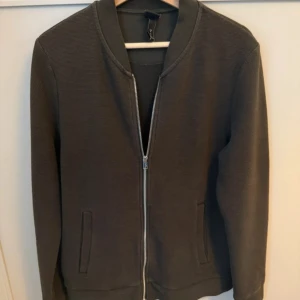 Oliv bomberjacka i medium - Stilren oliv bomberjacka med dragkedja framtill och två fickor. Jackan har ribbade muddar vid krage, ärmslut och nederkant. Perfekt för dig som gillar en clean look och enkel design. 