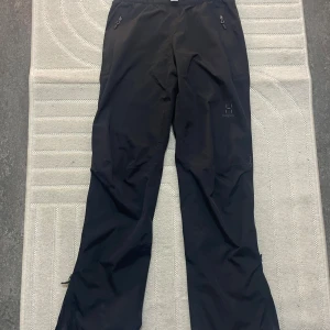 Svarta täckbyxor från Haglöfs - Svarta täckbyxor från Haglöfs med dragkedjor vid fickorna och justerbara benslut. Perfekta för utomhusaktiviteter under kyliga dagar. Byxorna har en enkel, rak passform och är tillverkade i slitstarkt syntetmaterial.