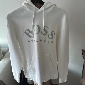 Vit hoodie från Hugo Boss - Snygg vit hoodie från Hugo Boss med stor logga på bröstet. Tröjan har huva med snörning och en klassisk känguruficka framtill. Mjuk bomullskvalitet och långärmad modell, köpt för 2000kr på Johnells Linköping