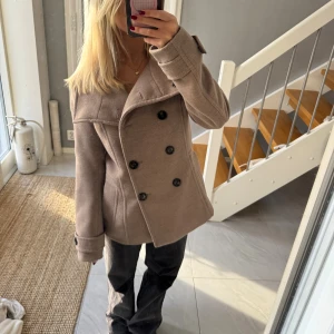 Beige kappa - Säljer en beige kappa med bred krage och dubbelknäppning framtill. Jackan har svarta knappar och snygga detaljer vid ärmsluten. Modellen är lite loose och passar perfekt till hösten. Materialet känns mjukt och har en ullig look.