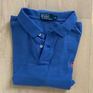 Blå pikétröja från Polo Ralph Lauren - Blå pikétröja från Polo Ralph Lauren i storlek XL. Klassisk krage, korta ärmar och två vita knappar framtill. Tröjan har den ikoniska röda Polo-loggan broderad på bröstet. Materialet är mjuk bomull som känns skönt mot huden.