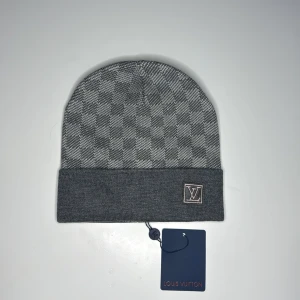 Lv mössa grå  - Snygg grå mössa från Louis Vuitton med klassiskt rutigt mönster och LV-logga framtill. Mössan har uppvikt kant och är tillverkad i mjukt material som passar perfekt för kyliga dagar.
