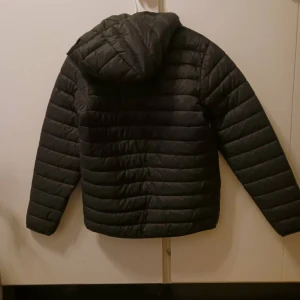 Svart pufferjacka från Ocean Works - Svart pufferjacka från Ocean Works i storlek 158/164. Jackan har huva, dragkedja framtill och två fickor med dragkedja. Quiltad design med horisontella sömmar och lätt vaddering. Perfekt för kyliga dagar och enkel att matcha med allt. Aldrig använt och inga skador.