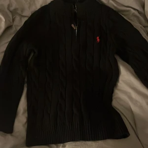 Svart stickad tröja Polo Ralph Lauren - Svart kabelstickad tröja från Polo Ralph Lauren med halv dragkedja och röd broderad logga på bröstet. Tröjan har hög krage, långa ärmar och ribbade muddar. Perfekt för dig som gillar klassisk och stilren design.