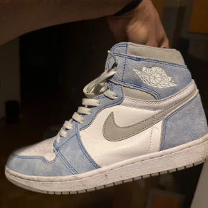 Nike Air Jordan 1 High blå/vit - Säljer ett par Nike Air Jordan 1 High med snygga blå mockadetaljer och vita läderpaneler. Skorna har grå swoosh och klassisk Air Jordan-logga på sidan. Perfekta för dig som vill sticka ut med en ikonisk sneaker i fräscha färger.