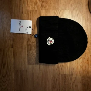 Svart ribbad Moncler mössa - Svart ribbstickad mössa från Moncler med klassisk uppvikt kant och ikonisk Moncler-logga framtill. Snygg och enkel design som passar till allt, tillverkad i mjukt stickat material för extra komfort. Perfekt för kalla dagar och en stilren look.
