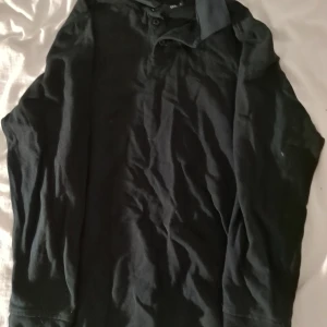 Svart långärmad pikétröja - Svart pikétröja med lång ärm och klassisk krage. Tröjan har knappar vid halsen och är gjord i mjukt bomullsmaterial. Perfekt för dig som gillar en enkel och stilren look. Passar dig som vill ha en basic tröja med lite mer edge.