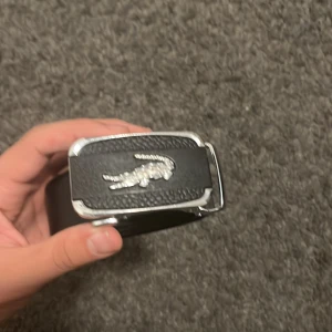 Svart Lacoste bälte i skinn - Snyggt svart bälte från Lacoste i skinn med silverfärgat spänne och den klassiska krokodilloggan framtill. Bältet har en stilren och clean design som passar till många outfits. Perfekt accessoar för dig som gillar att lyfta looken med en ikonisk detalj.