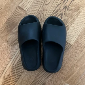Svarta Adidas Yeezy Slides - Säljer ett par svarta Adidas Yeezy Slides med chunky, räfflad sula och minimalistisk design. Tillverkade i syntetmaterial för en mjuk och bekväm känsla. Perfekta för chill dagar och enkel att glida i med öppen tå och stilren look.