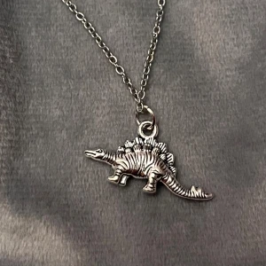 Halsband Dinosaurie Smycke Silver Imitation  - Så fint halsband med en berlock av en dinosaurie, en stegosaurus i detta fallet.  Halsbandet är justerbart 40-45 cm och är i silverimitation. Berlocken är ca 2,5 x 1 cm. 