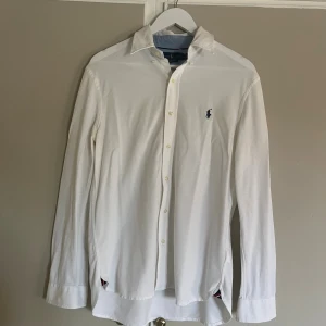 Vit långärmad skjorta från Ralph Lauren - Vit skjorta från Ralph Lauren i storlek M, tillverkad i lätt mesh-bomull. Klassisk krage, knappar framtill och på ärmslut, samt blått broderat logomärke på bröstet. Snygga blå detaljer på insidan av kragen och ärmsluten ger extra stilpoäng.
