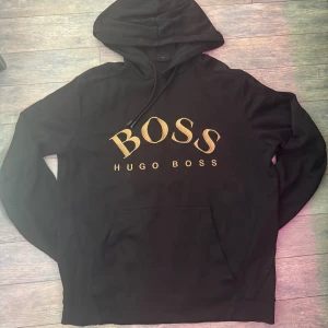 Svart hoodie från Hugo Boss (M) - Svart hoodie från Hugo Boss med stor guldfärgad BOSS-logga på bröstet. Klassisk modell med huva, snörning och känguruficka. Perfekt för dig som gillar stilrena streetwear-plagg med premiumkänsla. (Använt den ganska mycket, fast finns inge skador elle något), pris kan diskuteras