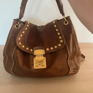 Brun axelväska från Miu Miu i skinn - Snygg brun axelväska från Miu Miu i mjukt skinn med guldfärgade nitar och låsdetalj framtill. Väskan har både handtag och axelrem, samt vita kontrastsömmar som ger en cool touch. Perfekt storlek för allt du behöver och riktigt trendig design.