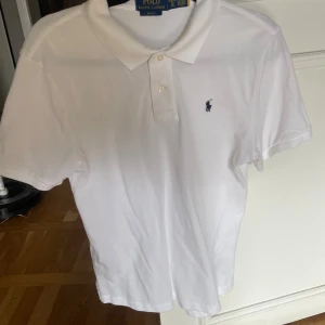 Vit pikétröja från Ralph Lauren - Klassisk vit pikétröja från Ralph Lauren i slim fit-modell. Tröjan har korta ärmar, krage med två knappar och den ikoniska broderade loggan på bröstet. Perfekt för en clean och stilren look. Tillverkad i mjuk bomull.