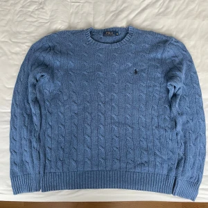 Blå kabelstickad tröja Polo Ralph Lauren - Kabelstickad blå tröja från Polo Ralph Lauren i bomull. Klassisk rund halsringning, ribbade muddar och ikonisk broderad logga på bröstet. Perfekt för lager-på-lager och snygg till jeans eller chinos.